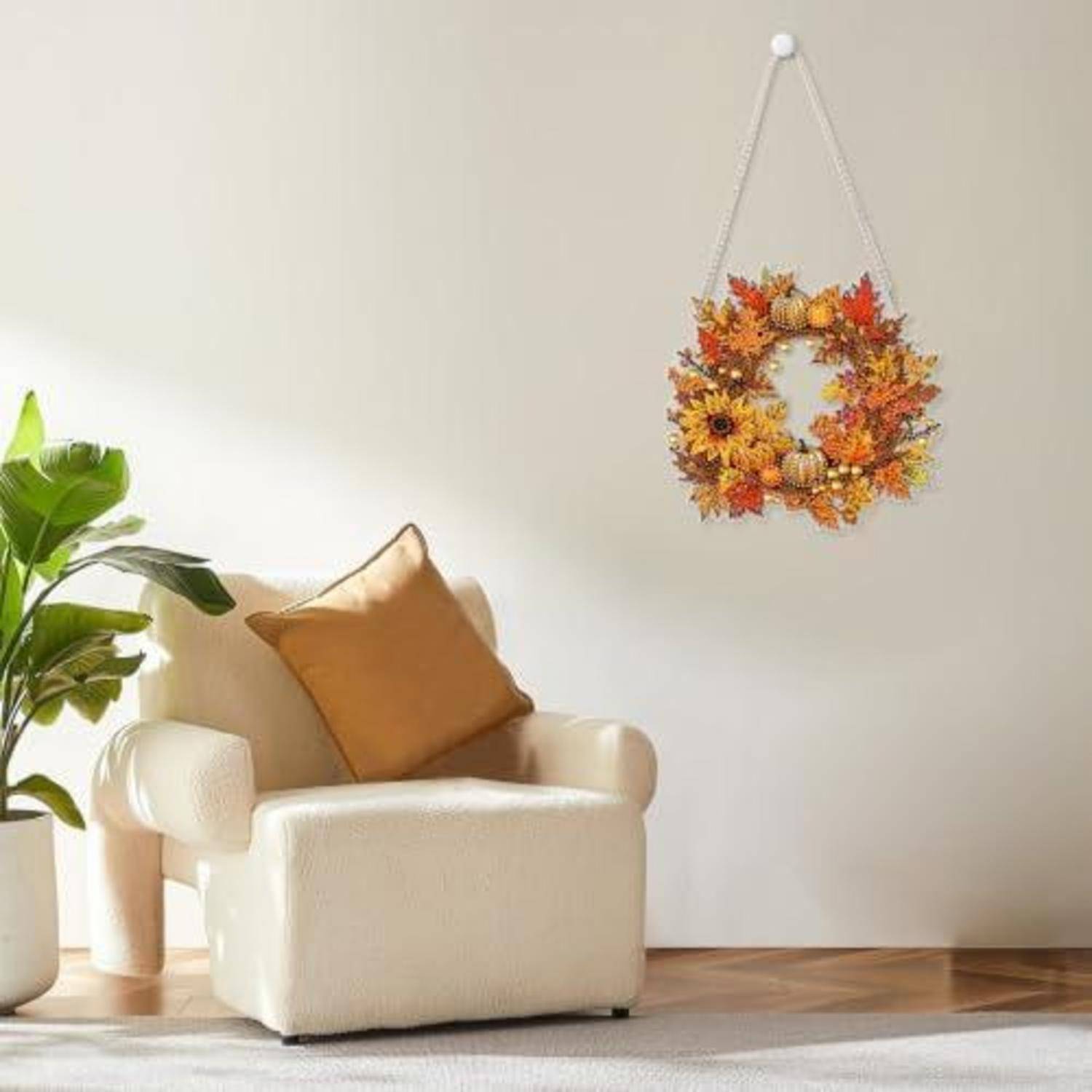 Gemütliches Wohnzimmer mit einem cremefarbenen Sessel, beigem Kissen, großer Pflanze und einem hängenden Herbstkranz an einer weißen Wand.