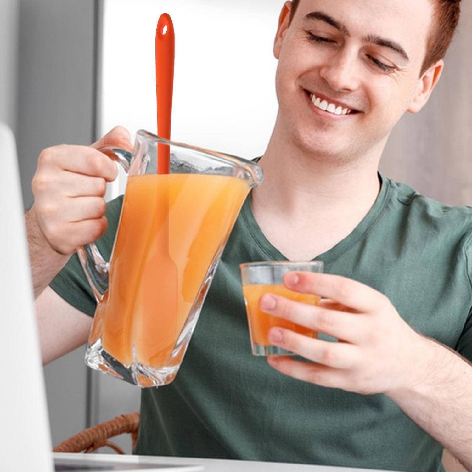 Lächelnder Mann gießt Orangensaft aus einem Krug in ein Glas, mit einem Löffel im Krug, in einer heimischen Küche.