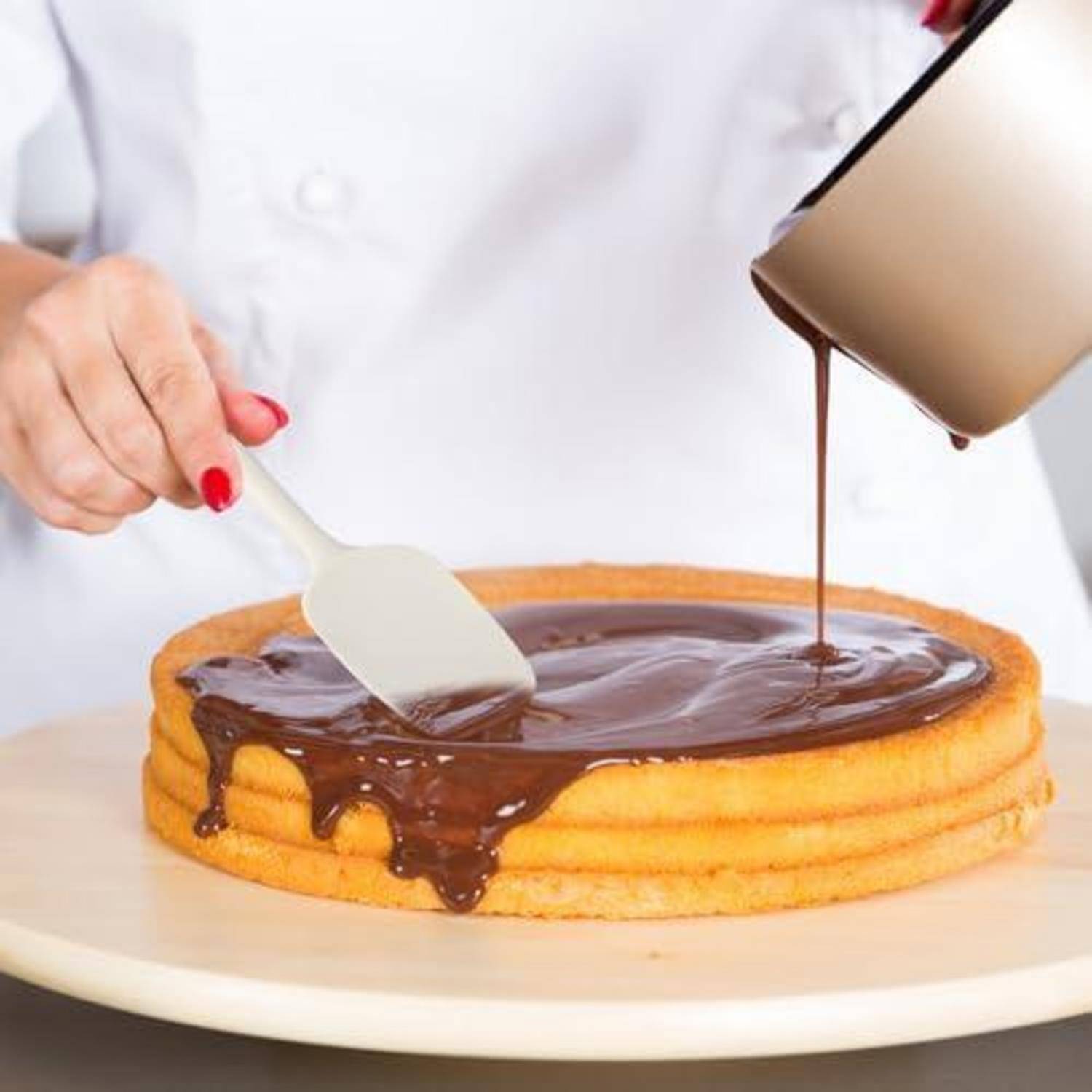 Eine Person verteilt Schokoladenglasur auf einem mehrschichtigen Kuchen, während sie gleichzeitig mehr aus einer Tasse auf die Oberseite gießt.