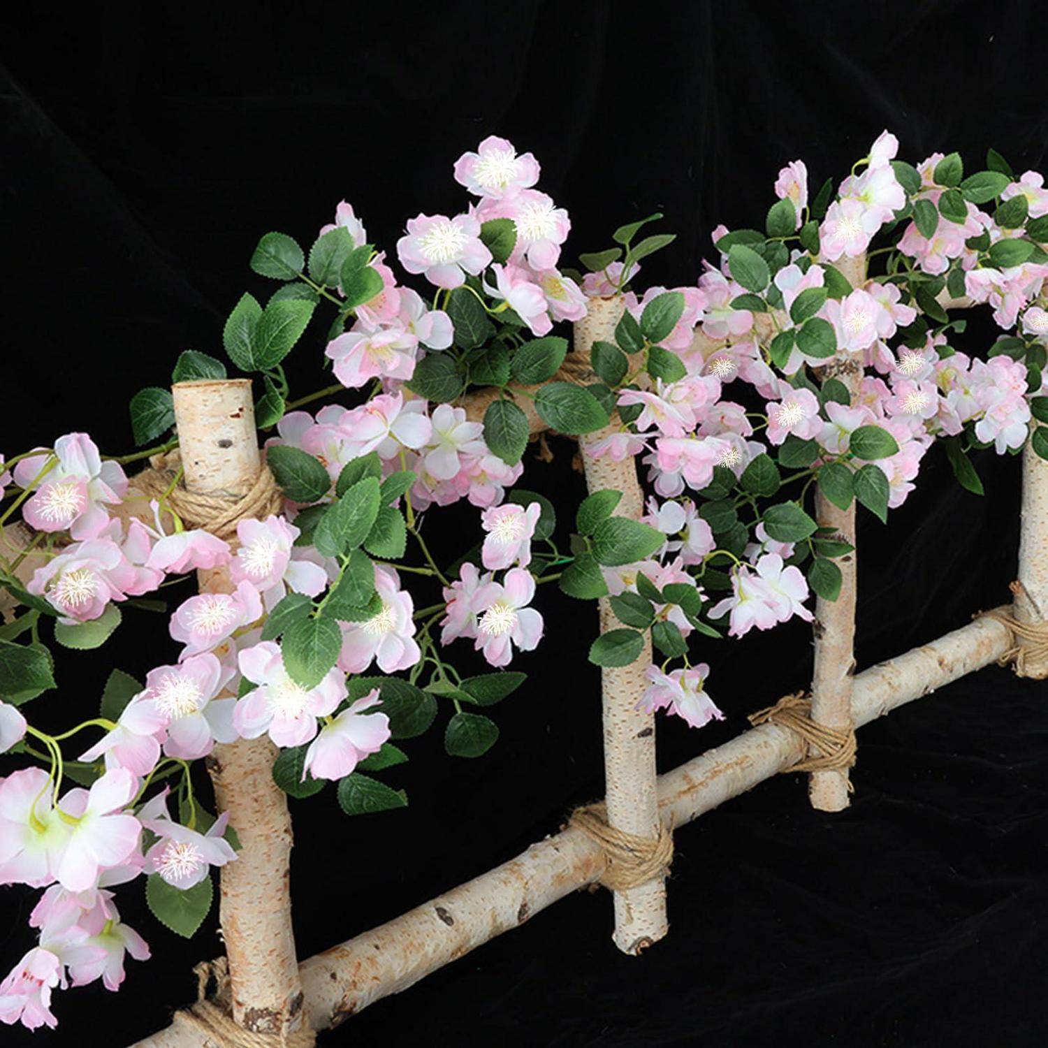 Dekorative Blütenarrangement aus hellrosa Blüten und grünen Blättern, angeordnet auf birkenähnlichen Ästen vor schwarzem Hintergrund.