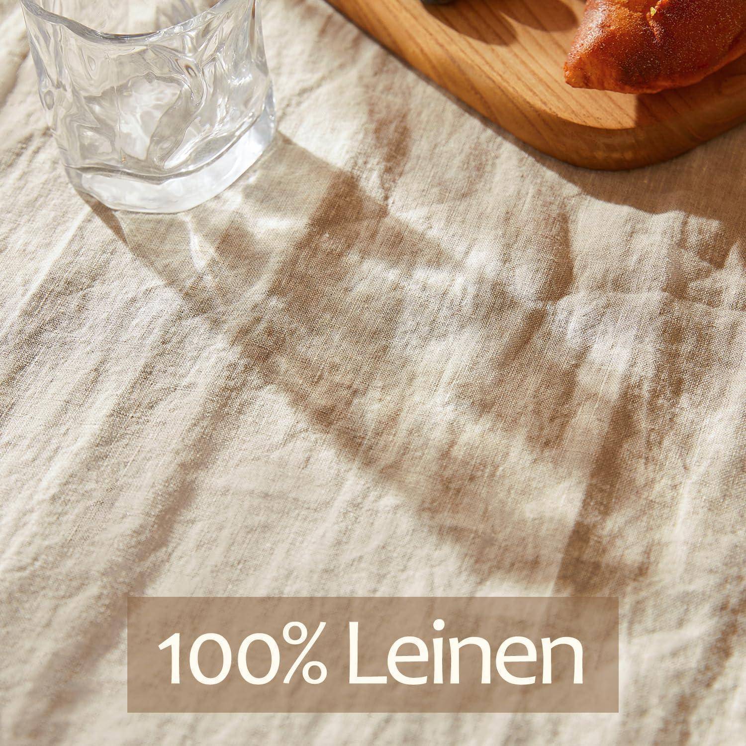Nahaufnahme eines Glases auf einem Leinengewebedecktuch mit Sonnenlichtschatten; der Text auf einem beigefarbenen Etikett lautet „100% Leinen".