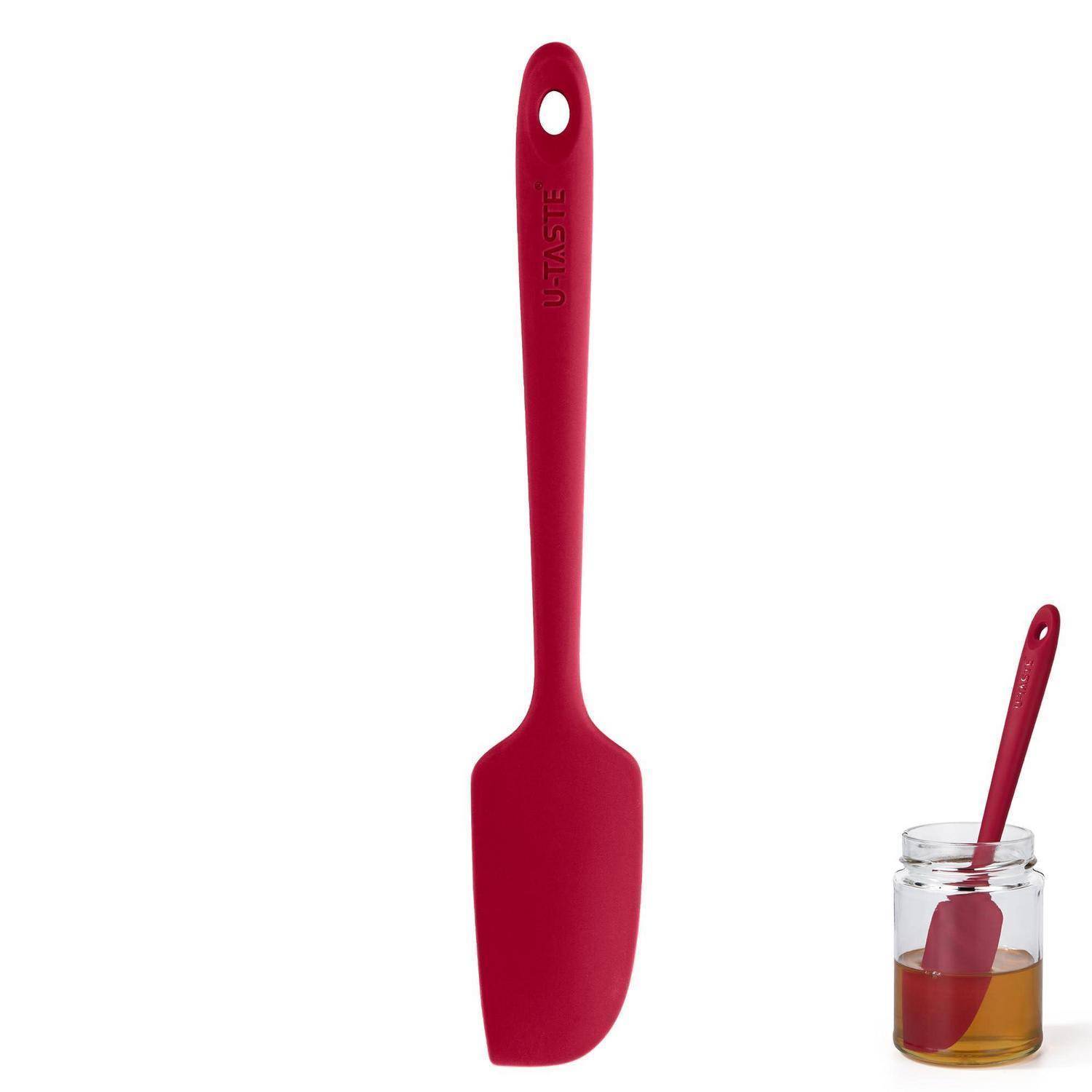Roter Silikonspatel mit Aufhängeloch, aufrecht auf weißem Hintergrund dargestellt, mit einem kleineren Spatel, der in einem Glasgefäß mit bernsteinfarbener Flüssigkeit ruht.