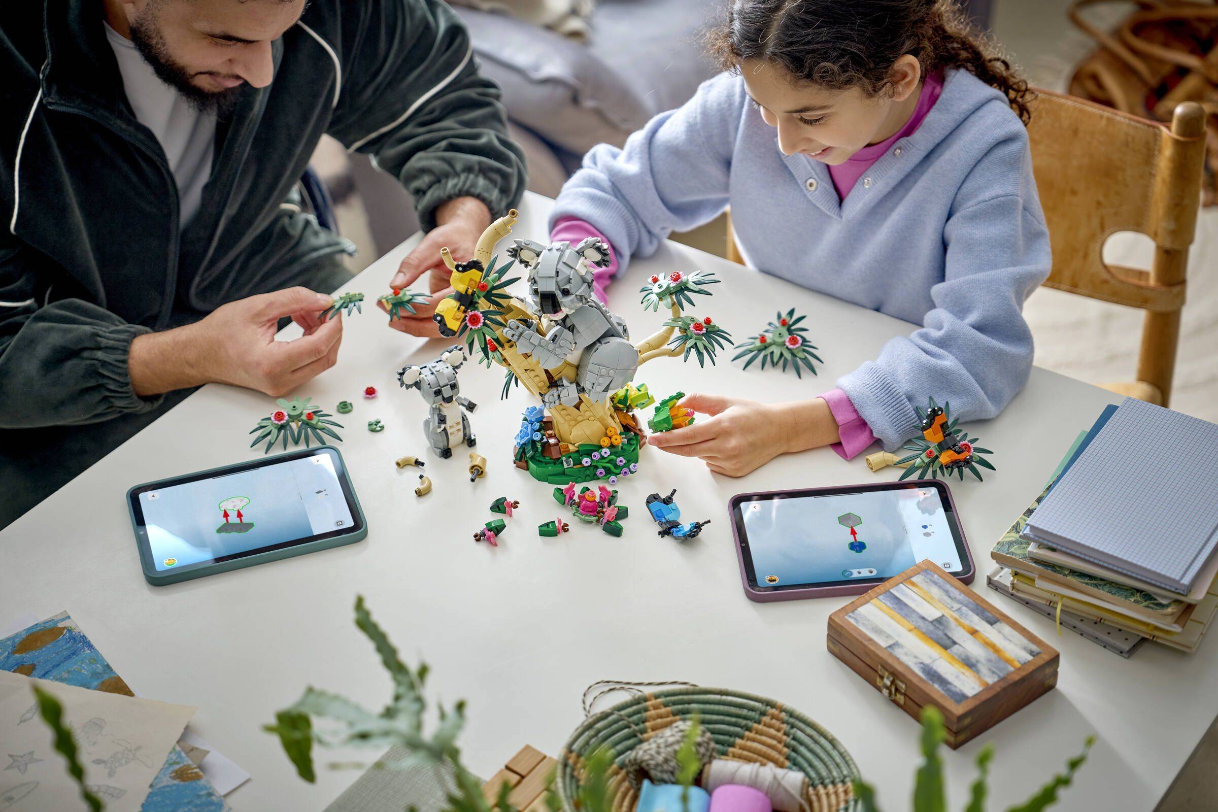 Ein Erwachsener und ein Kind bauen einen Spielzeugroboter aus bunten Bausteinen an einem Tisch, mit zwei Tablets in der Nähe und verstreuten Teilen um sie herum.