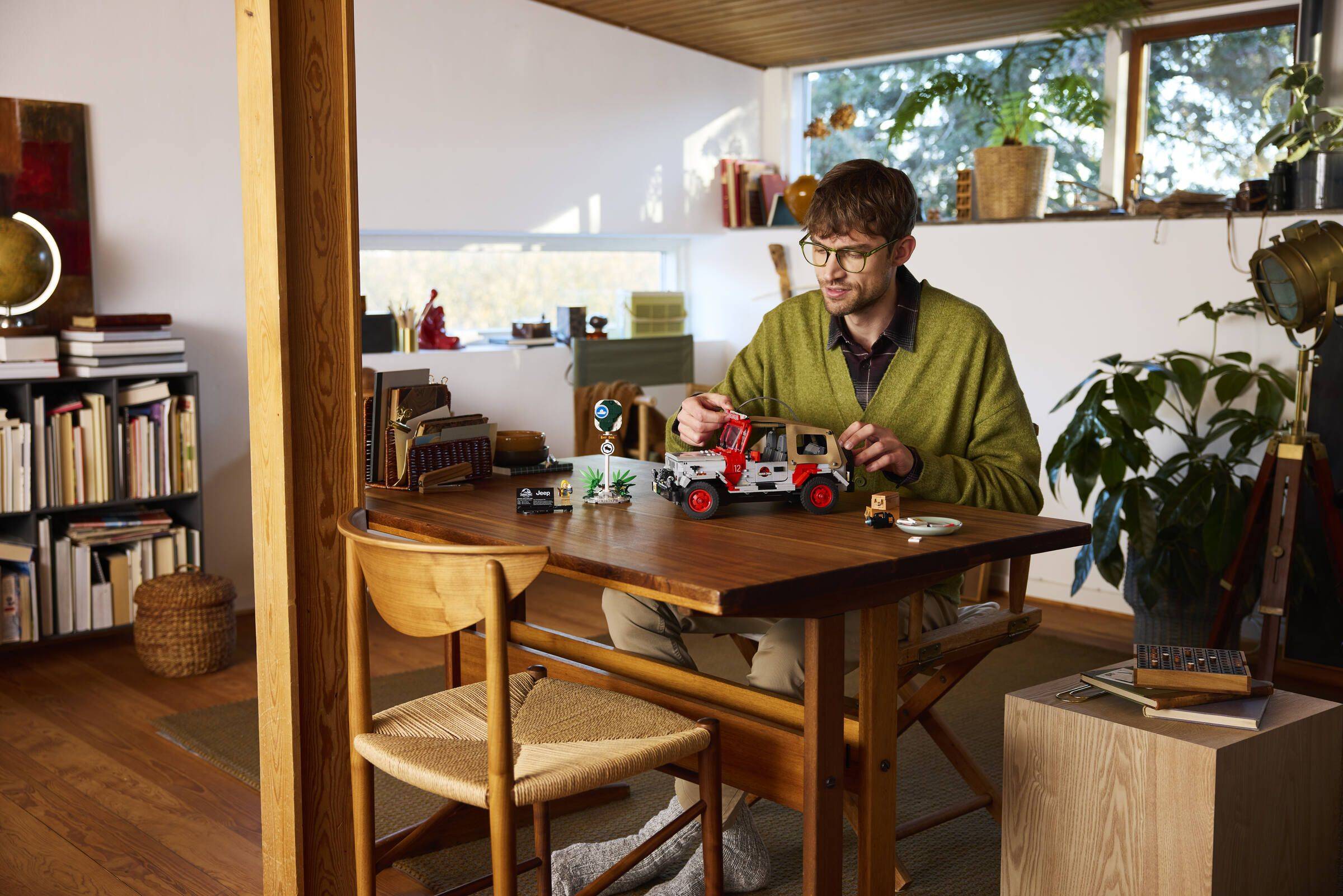 Ein Mann mit Brille montiert ein rotes Spielzeugfahrzeug an einem Holztisch in einem sonnigen Zimmer, umgeben von Bücherregalen und Zimmerpflanzen.