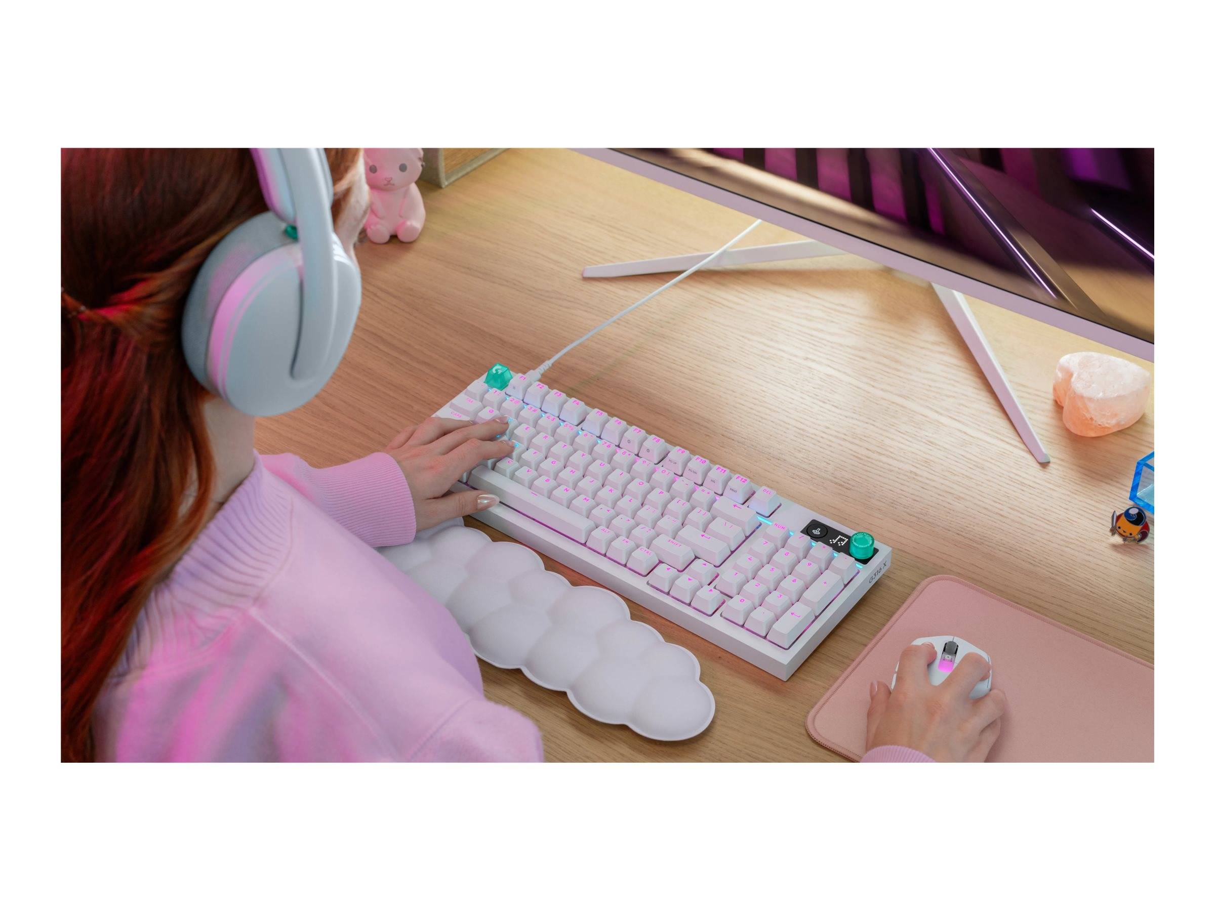 Person mit Kopfhörern, die eine rosa Maus und weiße Tastatur an einem Schreibtisch mit Monitor verwendet, Gaming-Setup oder Computer-Arbeitsbereich.