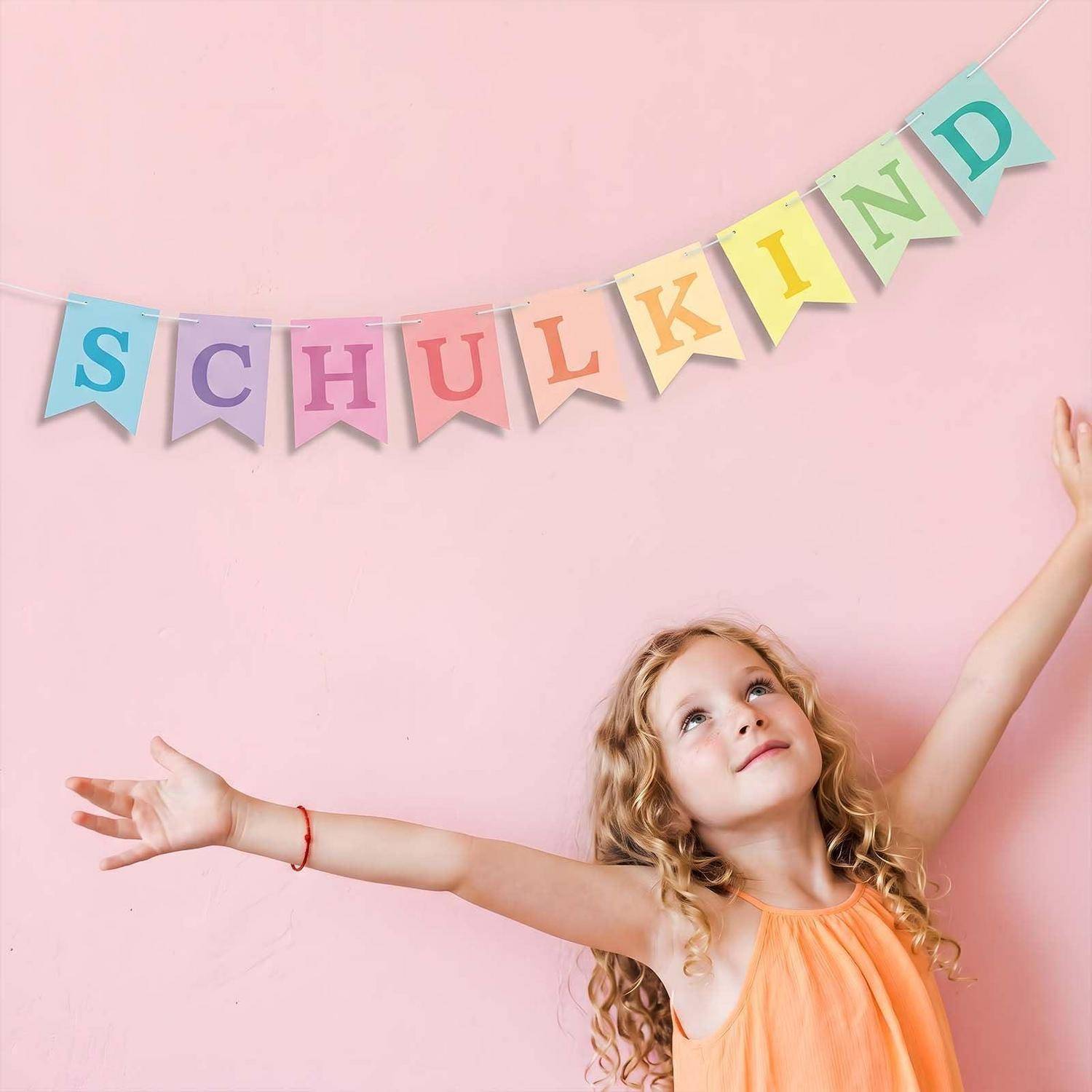 Junges Mädchen mit ausgestreckten Armen unter einem Banner mit der Aufschrift „SCHULKIND" vor einer rosa Wand, das den ersten Schultag feiert.
