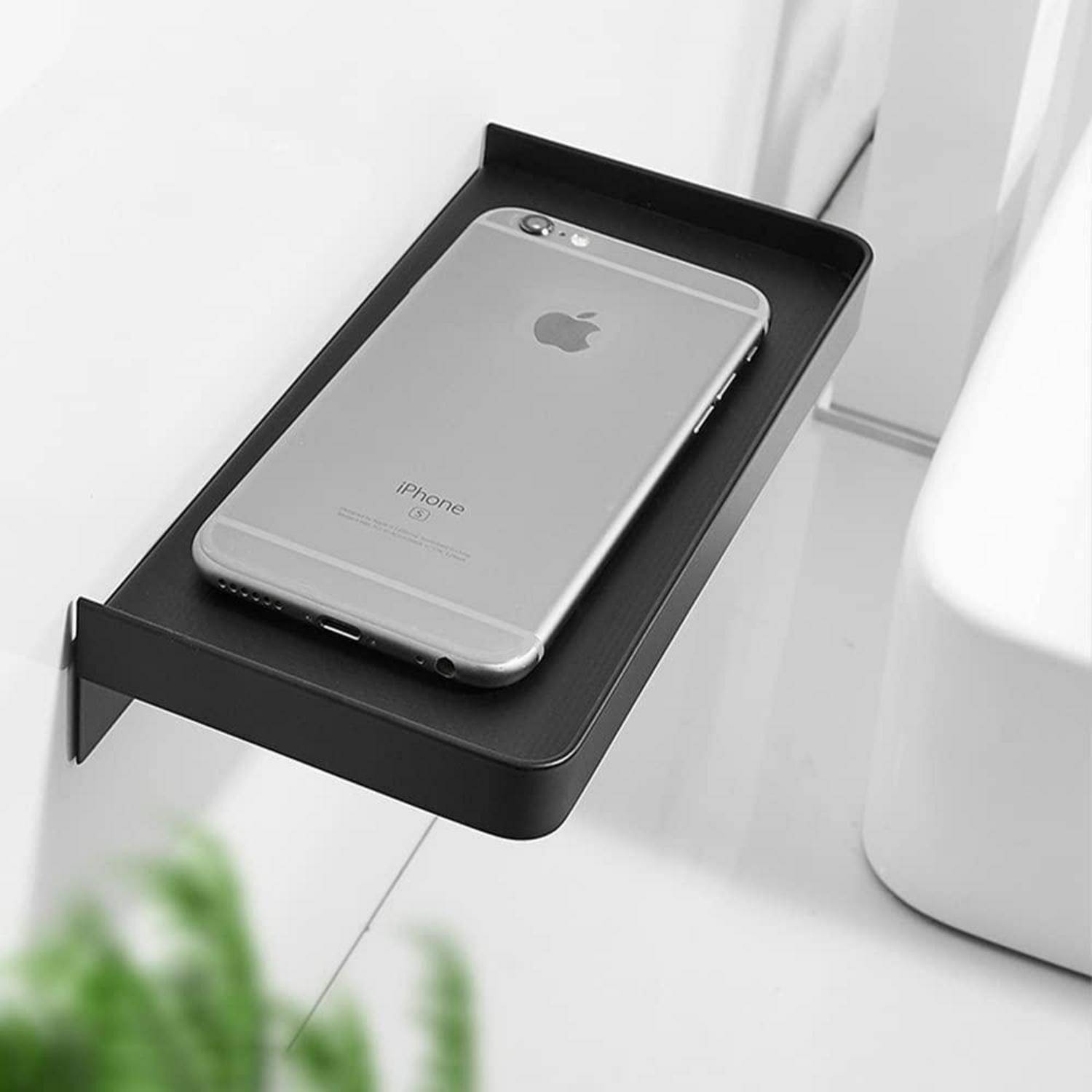 Ein silbernes iPhone liegt auf einem schwarzen, an der Wand befestigten Regal neben einem weißen Waschbecken, was auf eine kompakte Badezimmer-Aufbewahrungslösung hindeutet.