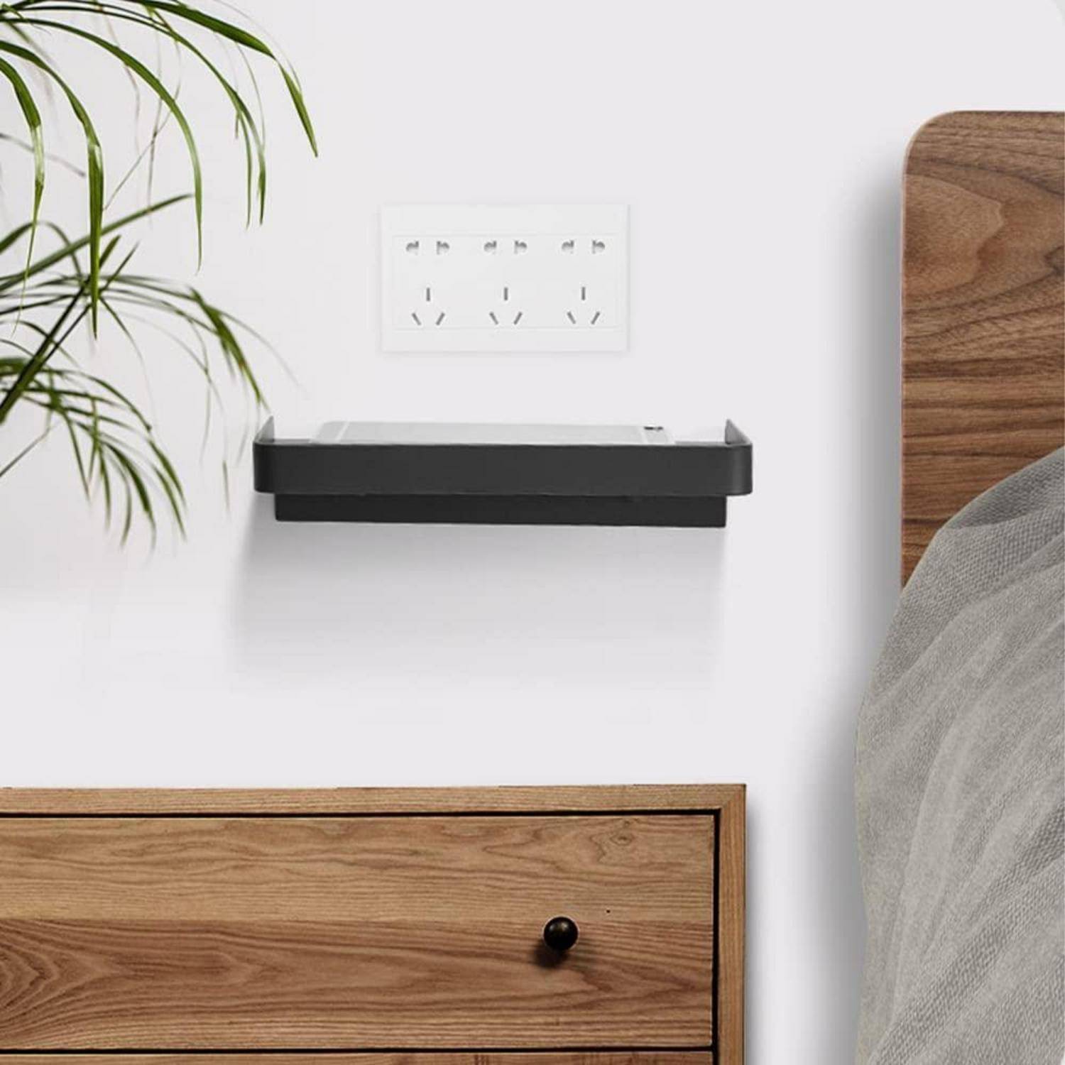 An der Wand montiertes schwarzes Regal unter einer Steckdose neben einem Bett mit Holzkopfteil und einer Pflanze auf der linken Seite.