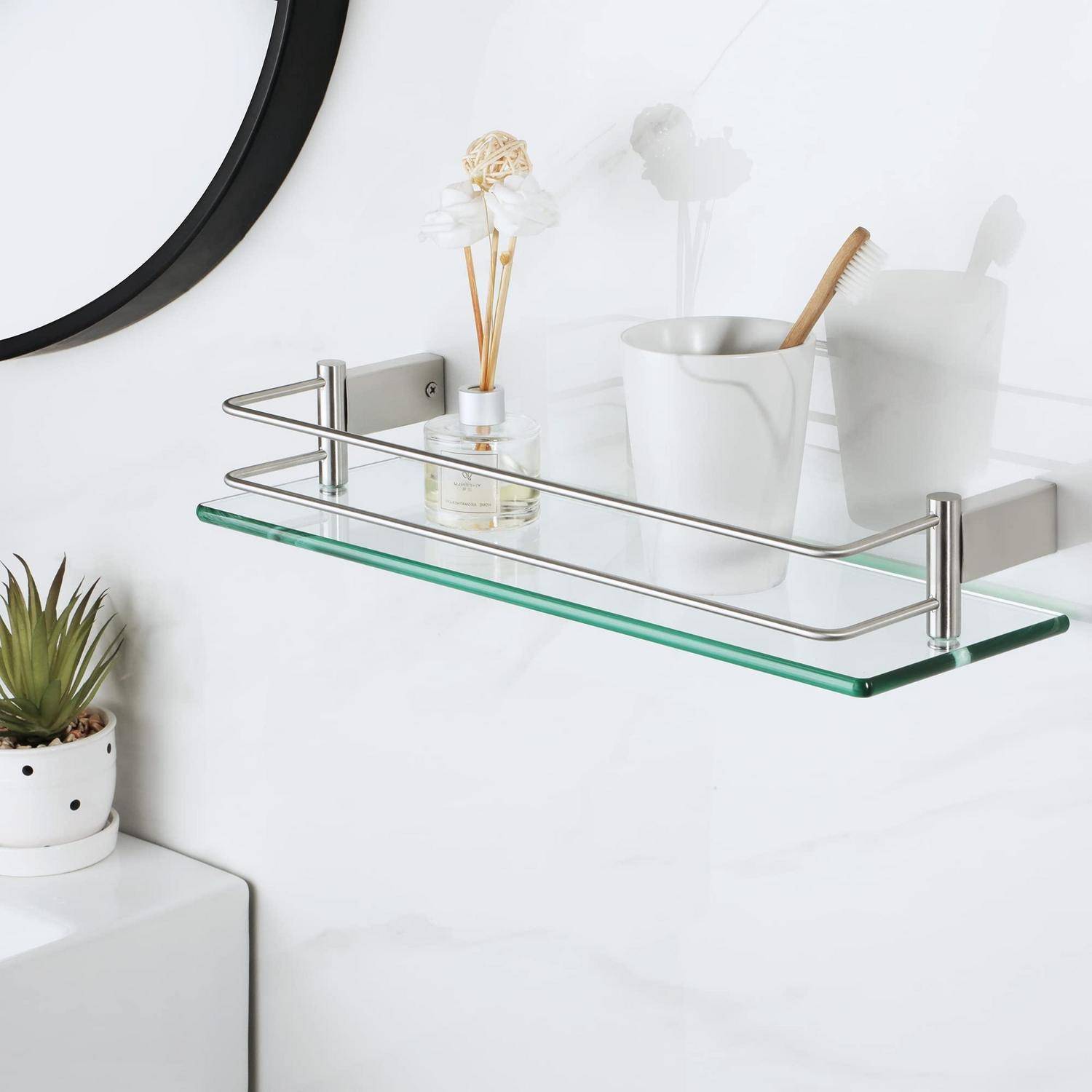 An der Wand montiertes Glassregal im Badezimmer mit einem Raumduft-Diffusor aus Rohr, einem Zahnbürstenbecher und kleiner Dekoration neben einem Spiegel und einer Topfpflanze.