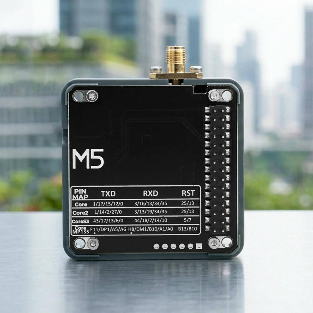 Schwarzes M5-Geräte-Modul mit Antennenstecker und beschrifteter Pinbelegung, auf einem Tisch mit unscharfem Stadthintergrund dargestellt.