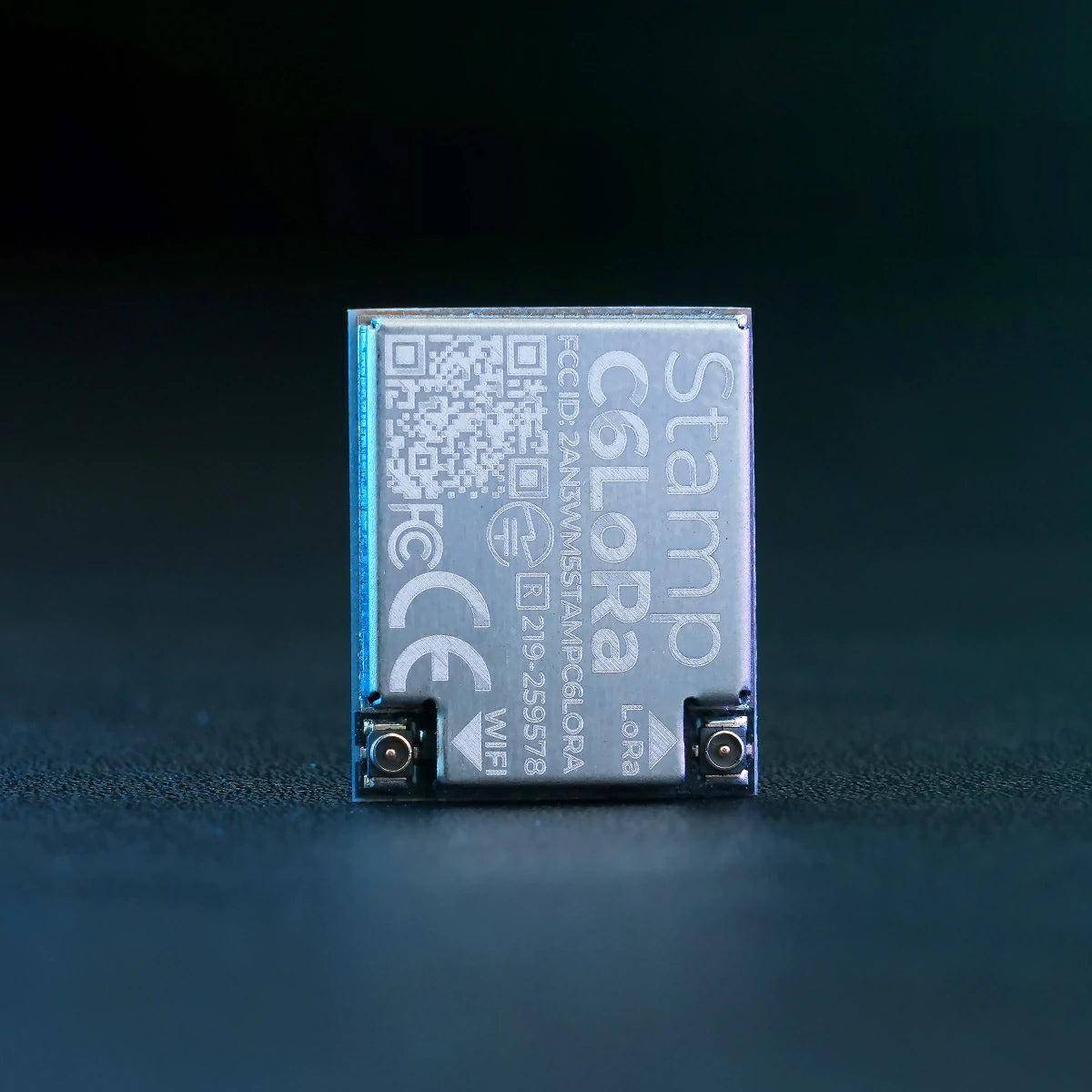 Ein kleines transparentes elektronisches Modul mit der Bezeichnung „Stamp Color" steht aufrecht auf einer dunklen Oberfläche und zeigt QR-Codes, Zertifizierungsmarken und zwei Metallkonnektoren.