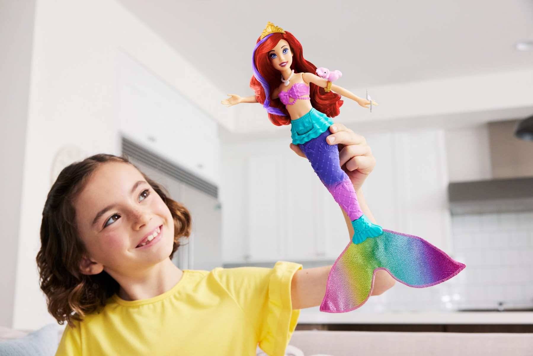 Lächelndes Mädchen in einem gelben Hemd, das eine bunte Meerjungfrauen-Puppe in einem hellen Wohnzimmer hochhält.