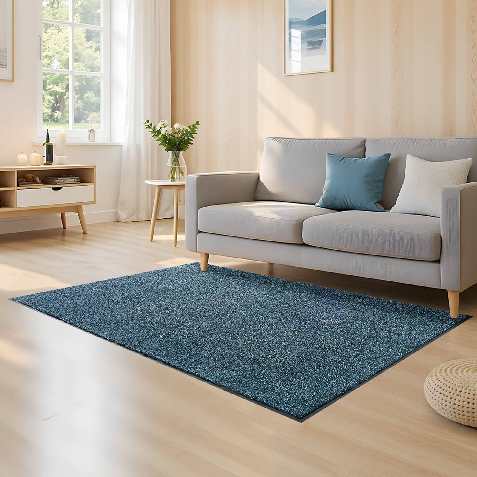 Helles Wohnzimmer mit grauem Sofa, blauem Teppich, kleinem Beistelltisch mit Blumen und Sonnenlicht durch ein Fenster.