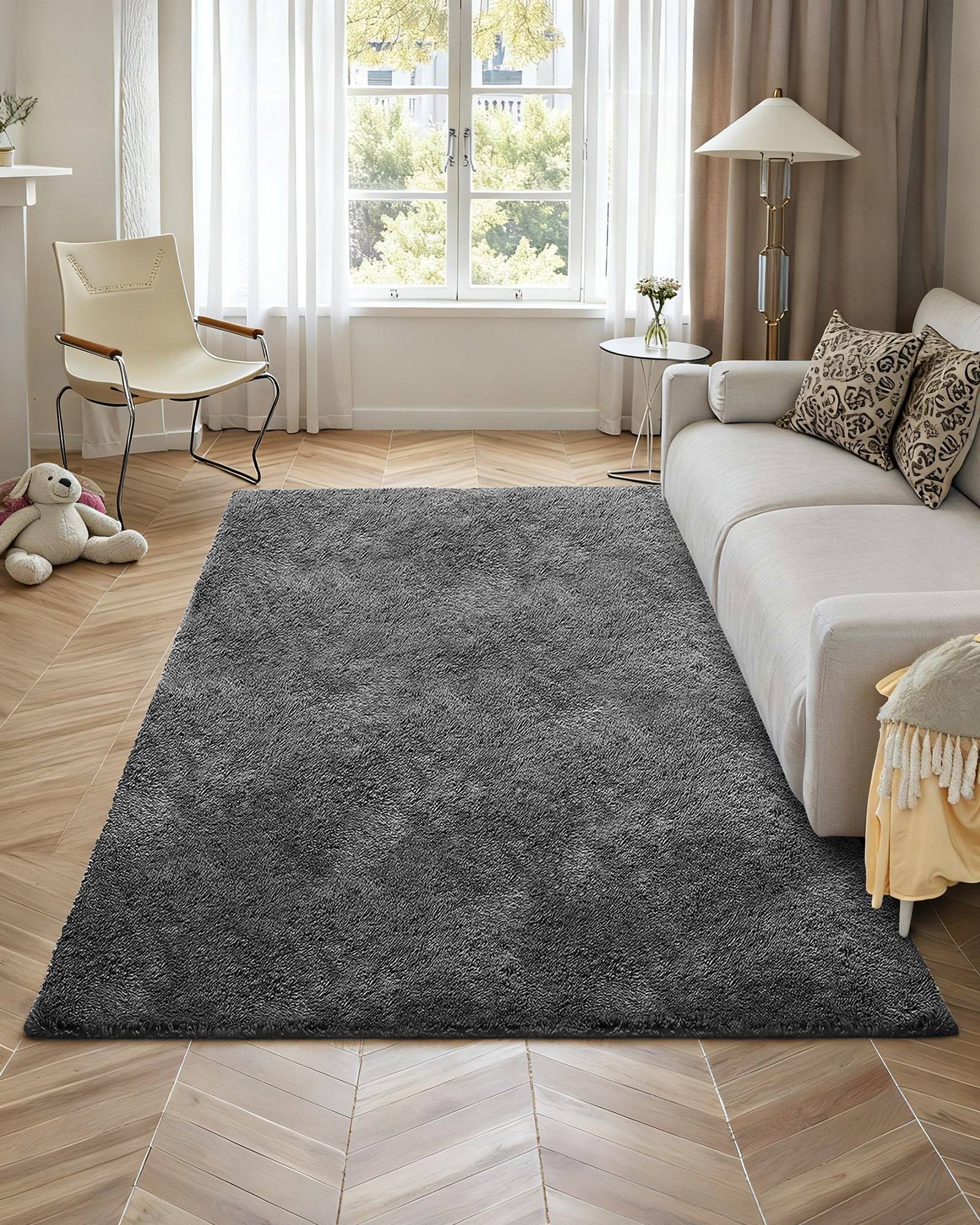 Ein gemütliches Wohnzimmer mit einem großen dunkelgrauen Teppich, beigem Sofa, Sessel, Lampe, Fenster und einem Kuscheltier-Kaninchen auf dem Boden.