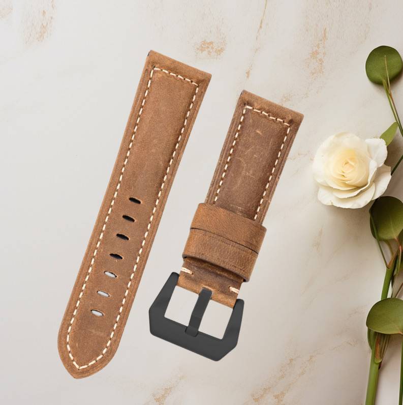 Braunes Lederarmband mit Nähten und schwarzer Schnalle, separat auf hellem Hintergrund mit einer weißen Blume und Blättern gezeigt.