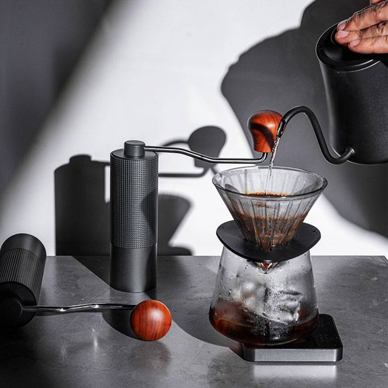 Pour-over-Kaffee wird in eine Glaskanne gegossen, wobei sich ein schwarzer Wasserkocher, eine Mühle und Kaffeemahlwerk-Werkzeuge auf einer Arbeitsplatte befinden.