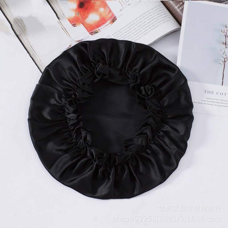Schwarze Satinhaube oder Duschkappe mit elastischem Rüschensaum, flach auf einer weißen Oberfläche neben aufgeschlagenen Büchern dargestellt.