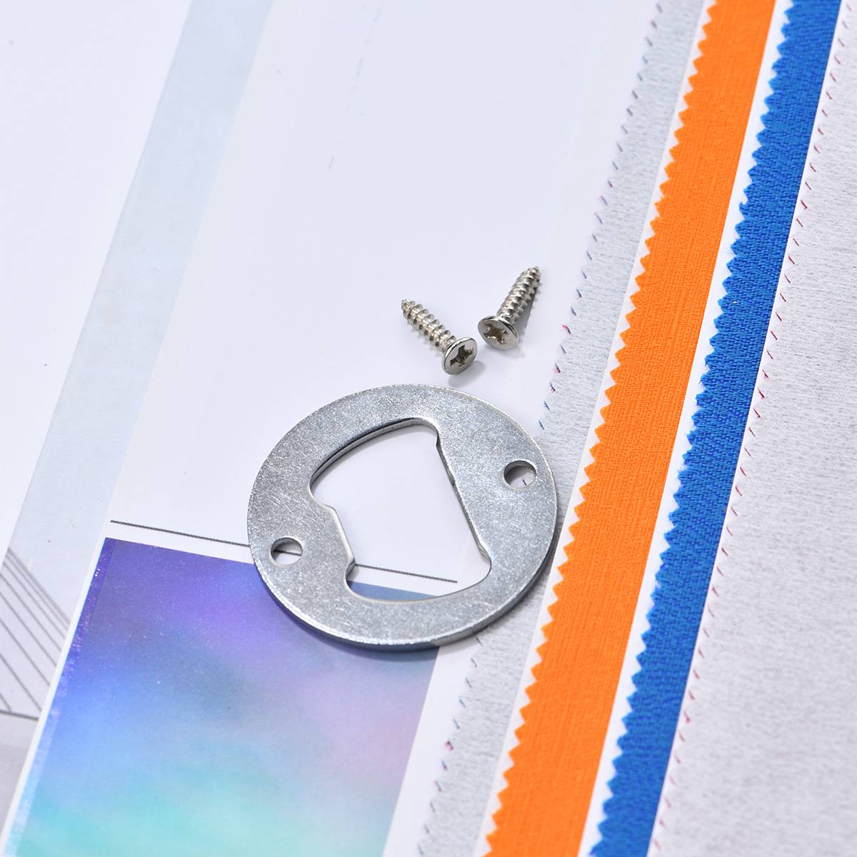 Metallischer Flaschenöffner-Ring und zwei Schrauben auf einem weißen Stoffmusterbuch mit orangefarbenen und blauen Randstreifen.