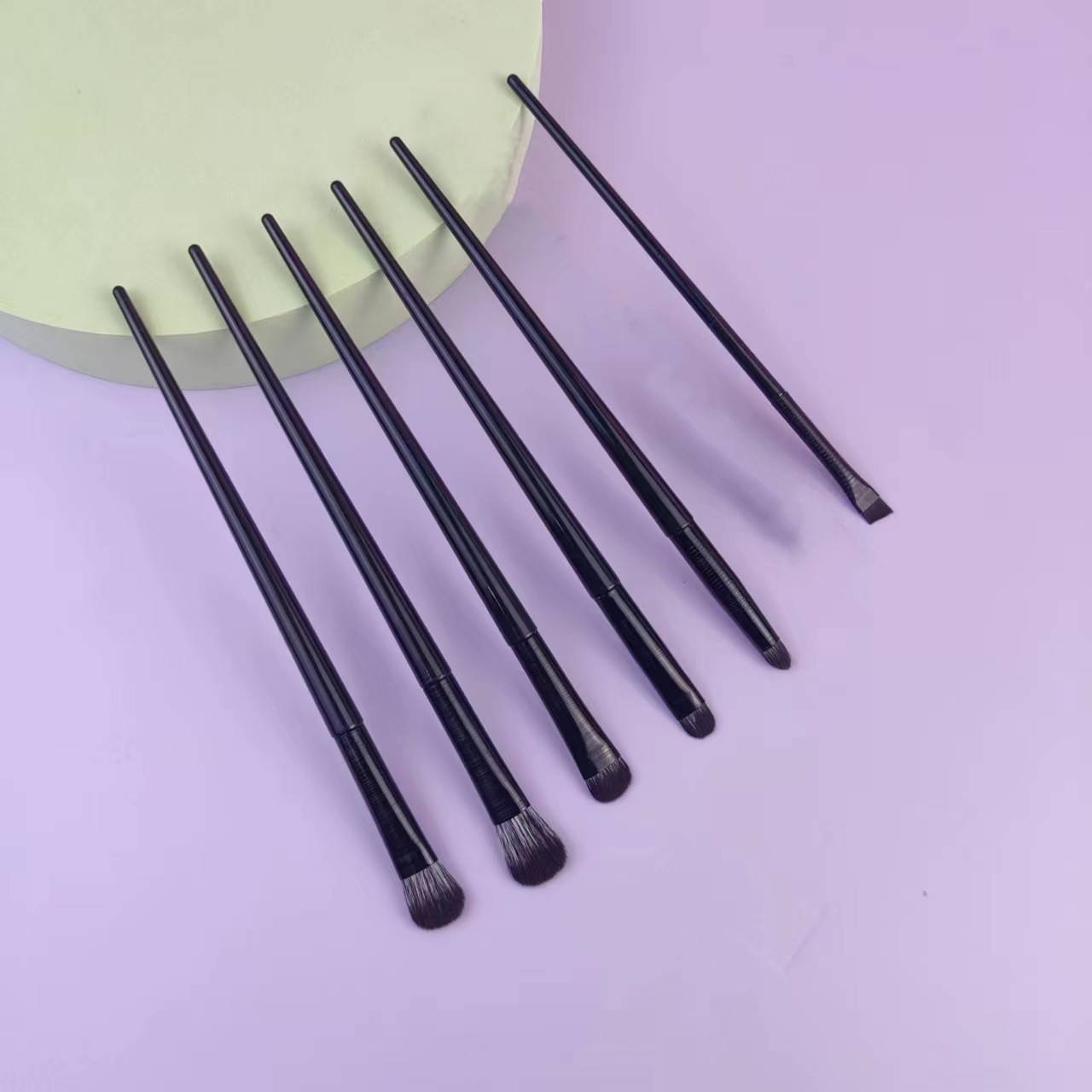 Sechs schwarze Makeup-Pinsel mit unterschiedlichen Spitzen, die auf einer pastellrosa Oberfläche neben einem blassen kreisförmigen Objekt angeordnet sind.