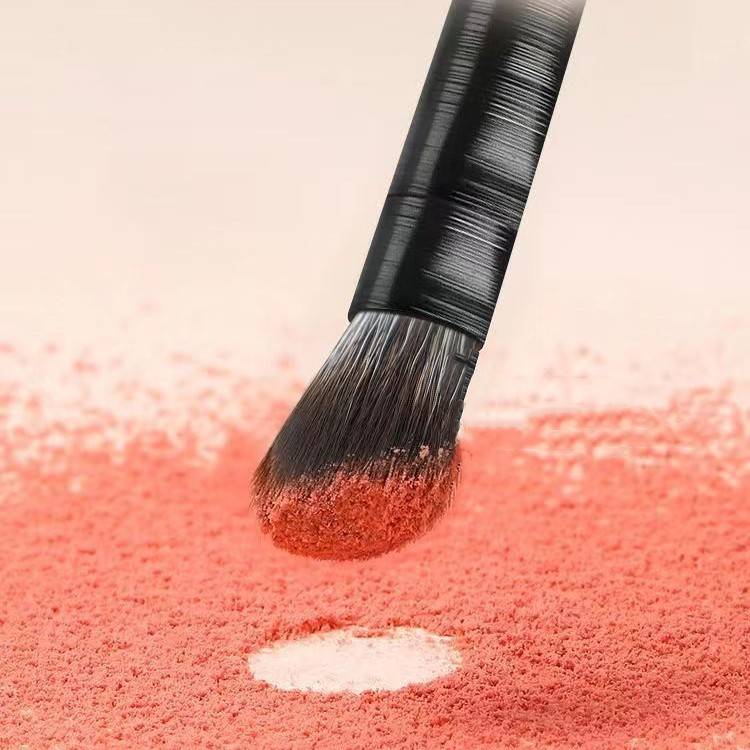 Ein Make-up-Pinsel trägt korallrosa Puder auf und verblendet ihn, wobei in der Mitte ein kleines weißes Highlight hinterlassen wird.