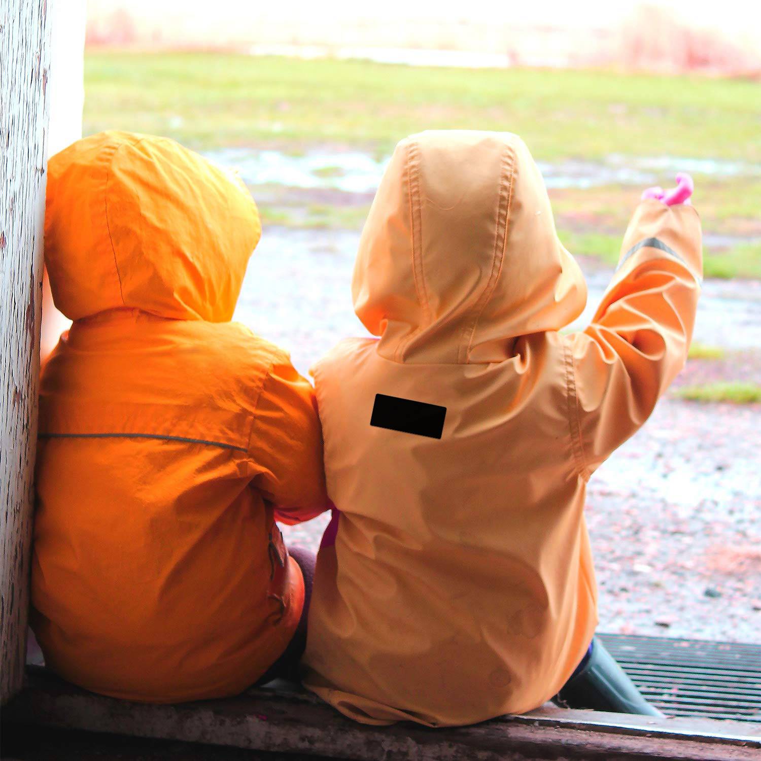 Zwei Kinder in orangefarbenen Regenmänteln sitzen nebeneinander in einer Türöffnung, wobei eines auf die nasse Außenwelt zeigt.