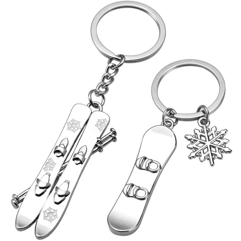 Zwei silberfarbene Ski-Schlüsselanhänger auf weißem Hintergrund, einer mit Skiern und Stöcken, der andere mit einem Ski und einem Schneeflocken-Charm.