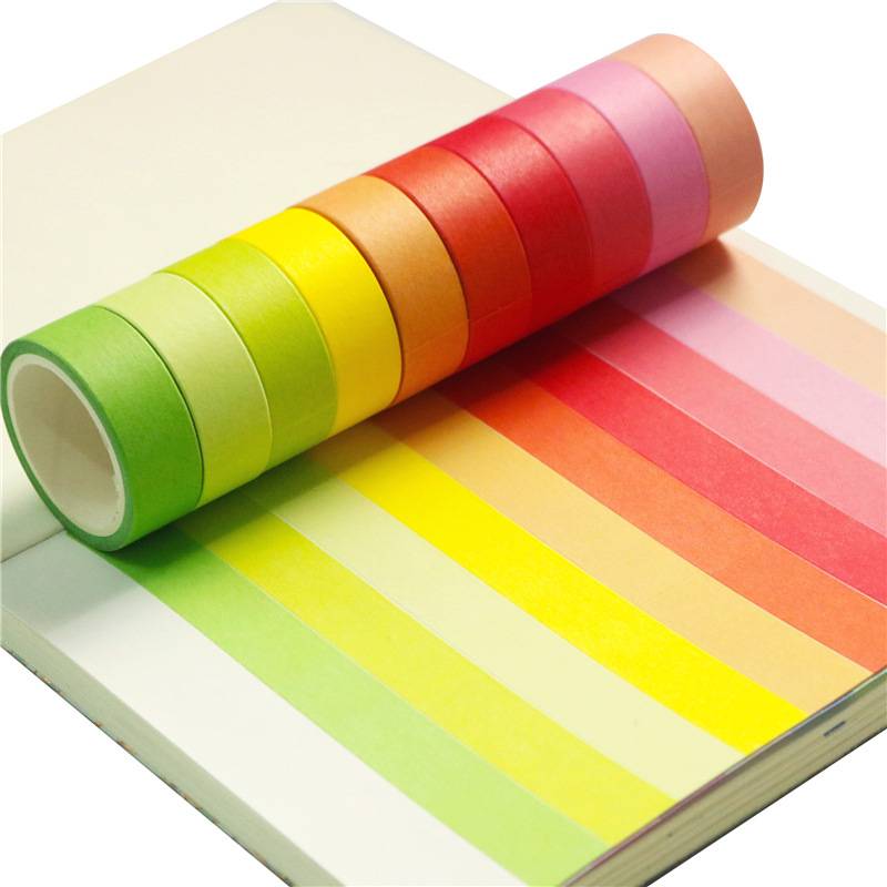 Regenbogenfarbige Klebeband-Rolle auf einem Notizbuch, mit parallel gestreiften Mustern, die fächerartig in Grün, Gelb, Orange, Rot und Rosa ausgebreitet sind.