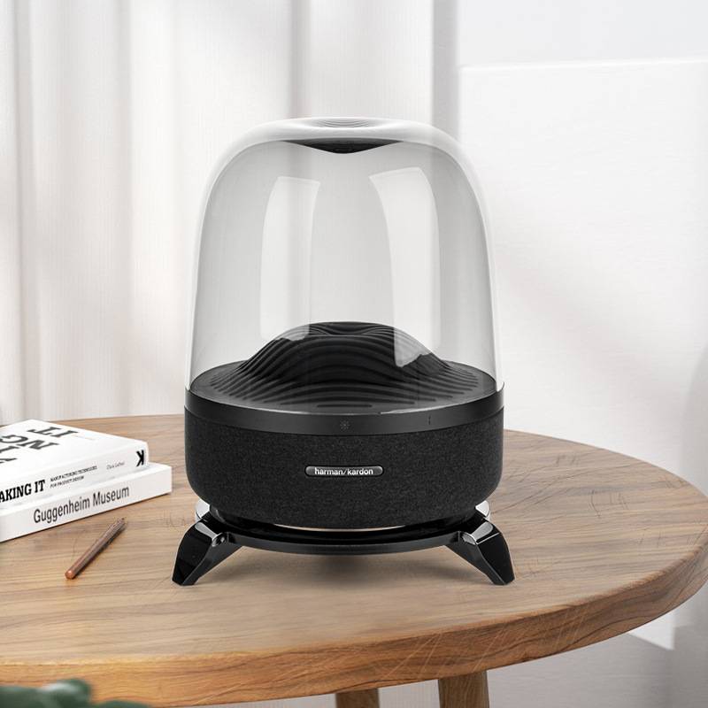 Schwarzer Harman Kardon Tischplattenlautsprecher mit einer transparenten Kuppelabdeckung auf einem hölzernen Beistelltisch neben Büchern und einem Bleistift.