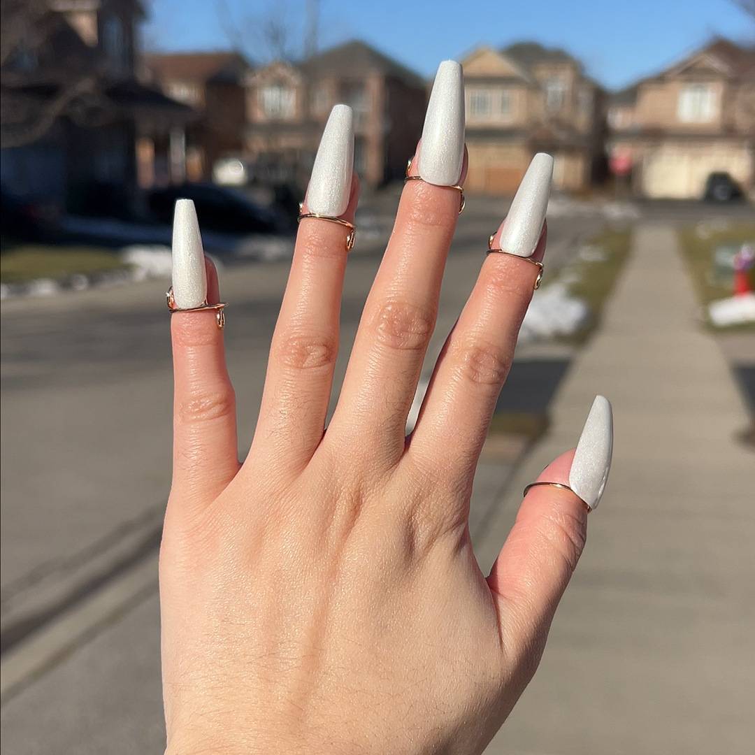 Hand mit langen weißen Stiletto-Acrylnägeln und goldenen Ringen, die an einem sonnigen Straße in einem Vorort im Freien hochgehalten wird.