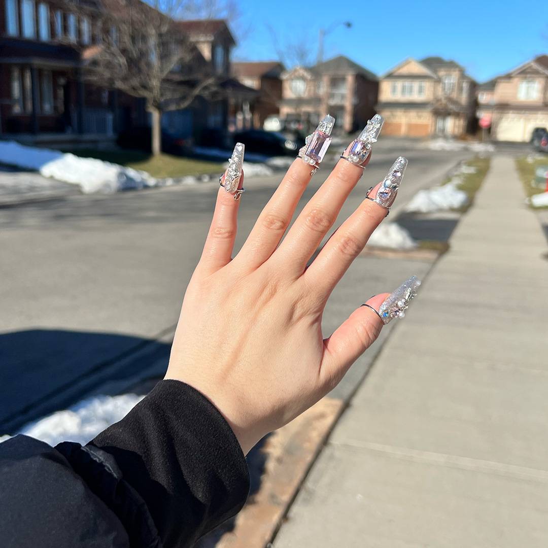 Hand mit erhobener Handfläche im Freien auf einer sonnigen Straße in einem Vorort, mit langen silbernen glitzernden Acrylnägeln.