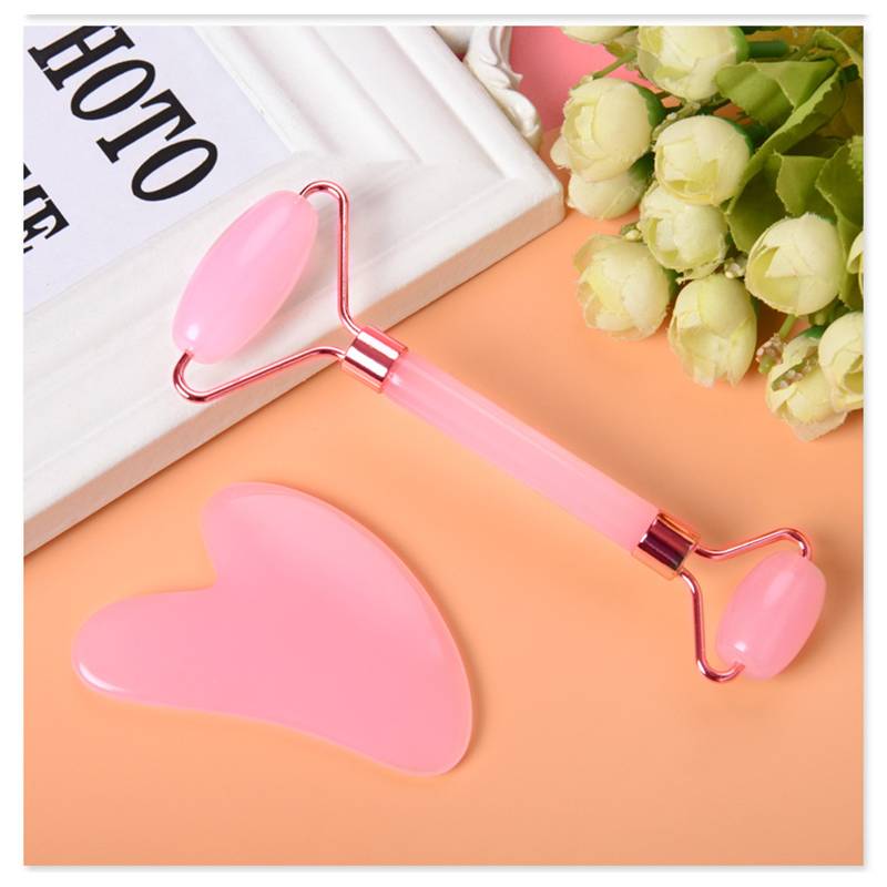 Rosa Gesichtsmassagerolle und Gua-Sha-Stein auf pfirsichfarbenem Hintergrund neben einem Buch und weißen Rosen.