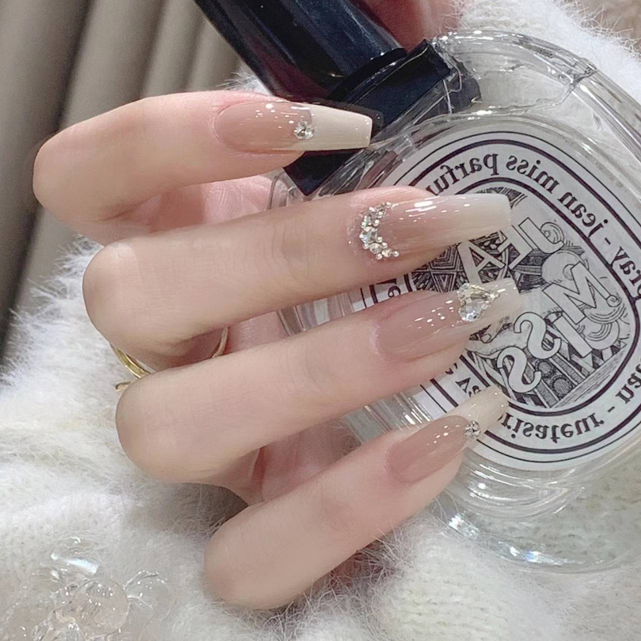 Nahaufnahme einer Hand mit langen, nackten Acrylnägeln, die mit Rhinestonen verziert sind und eine transparente Parfümflasche halten.