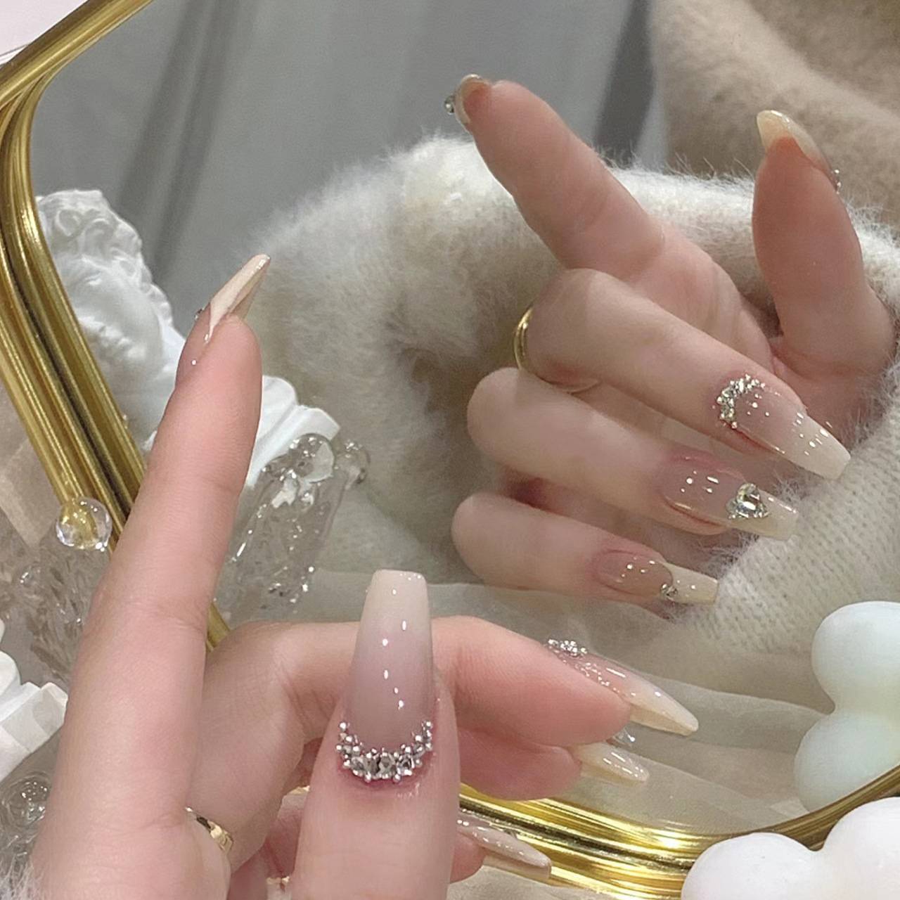 Nahaufnahme von Händen mit langen, nude-rosa manikürierten Nägeln, die mit Strass-Steinen verziert sind, gezeigt beim Halten eines vergoldeten Spiegels.