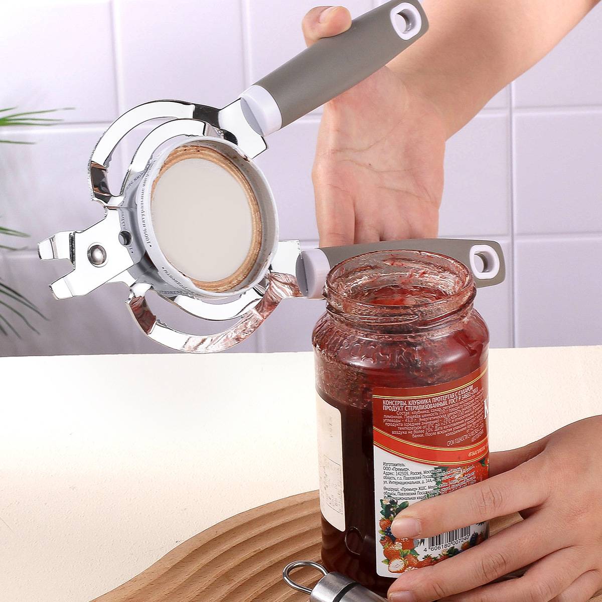Eine Hand benutzt ein Öffner-Werkzeug, um einen Marmeladenglas-Deckel auf einer Küchentheke zu greifen und aufzudrehen.