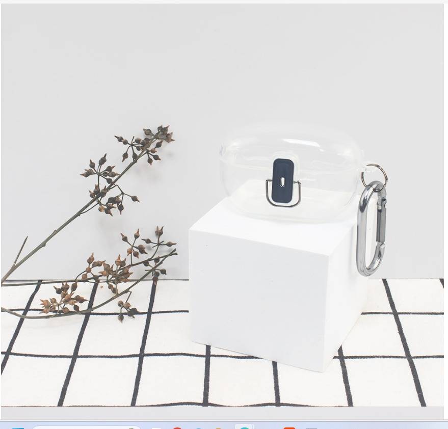 Transparentes AirPods-ähnliches Ladecase auf einem weißen Würfel, mit Karabinerhaken und getrockneter Pflanze auf einem schwarz karierten Stoffhintergrund.