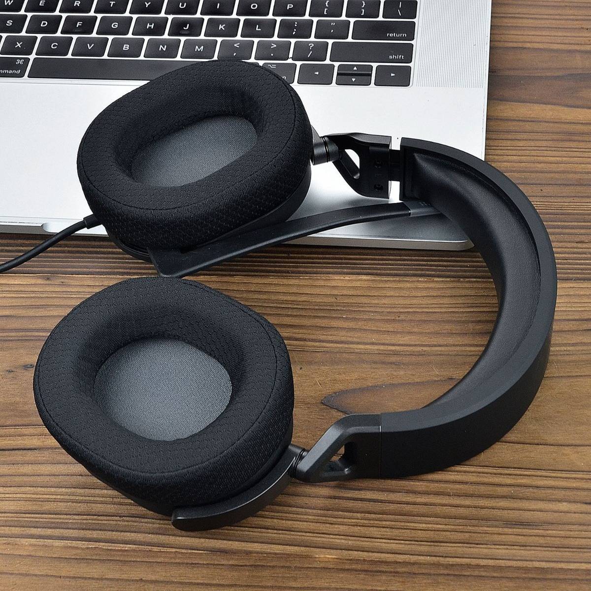 Schwarze Over-Ear-Kopfhörer ruhen auf einem Holzschreibtisch neben einer Laptop-Tastatur, wobei eine Ohrmuschel aufgeklappt ist.
