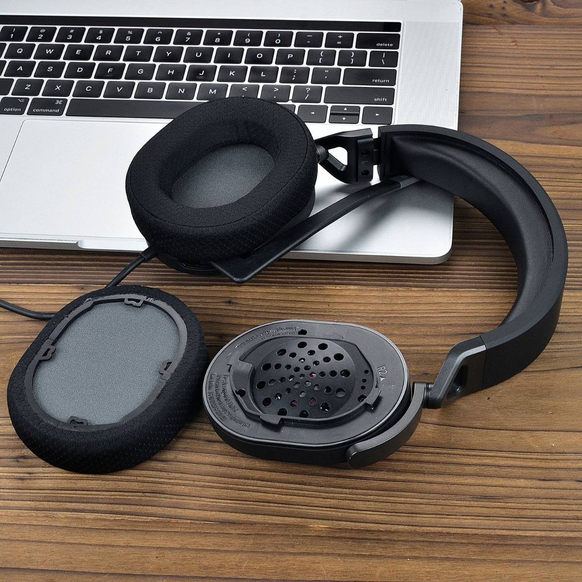 Schwarze Over-Ear-Kopfhörer sind auf einem Holzschreibtisch neben einem aufgeklappten Laptop abgebildet, wobei eine Ohrmuschel entfernt ist, um den Lautsprecher und das Polster zu zeigen.