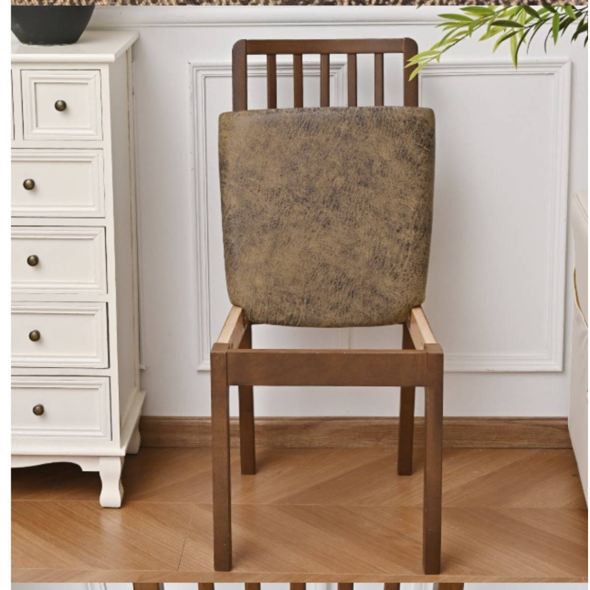 Holzstuhl mit melierter beigefarbener gepolsterter Rückenlehne, zentriert in einem hellen Zimmer neben einer weißen Kommode und Wandverkleidung.