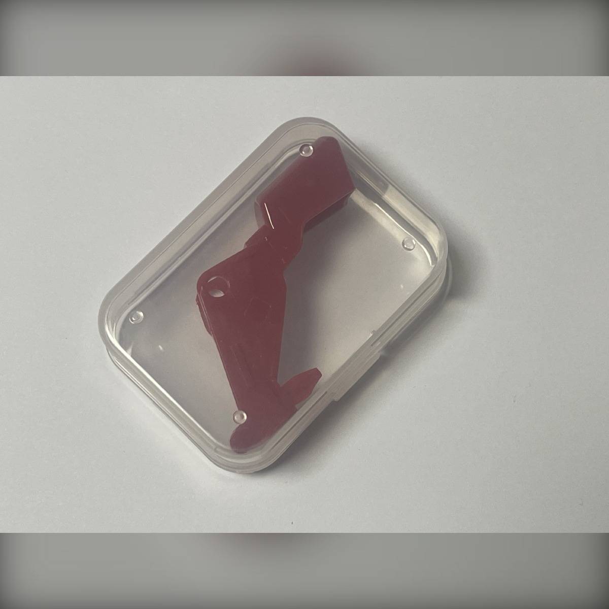 Rotes kunststoffteil mit auslöserform in einem durchsichtigen rechteckigen kunststoffgehäuse auf einer hellgrauen oberfläche.