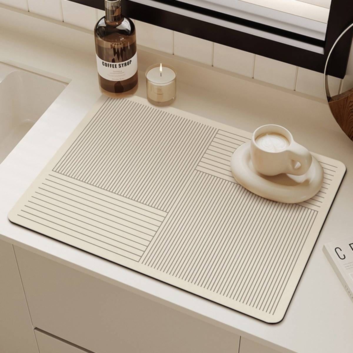 Eine beige gestreifte Schreibtischunterlage auf einer weißen Arbeitsplatte hält eine Kaffeetasse auf einer Untertasse, mit einer Kaffeesirupflasche und einer kleinen Kerze daneben.