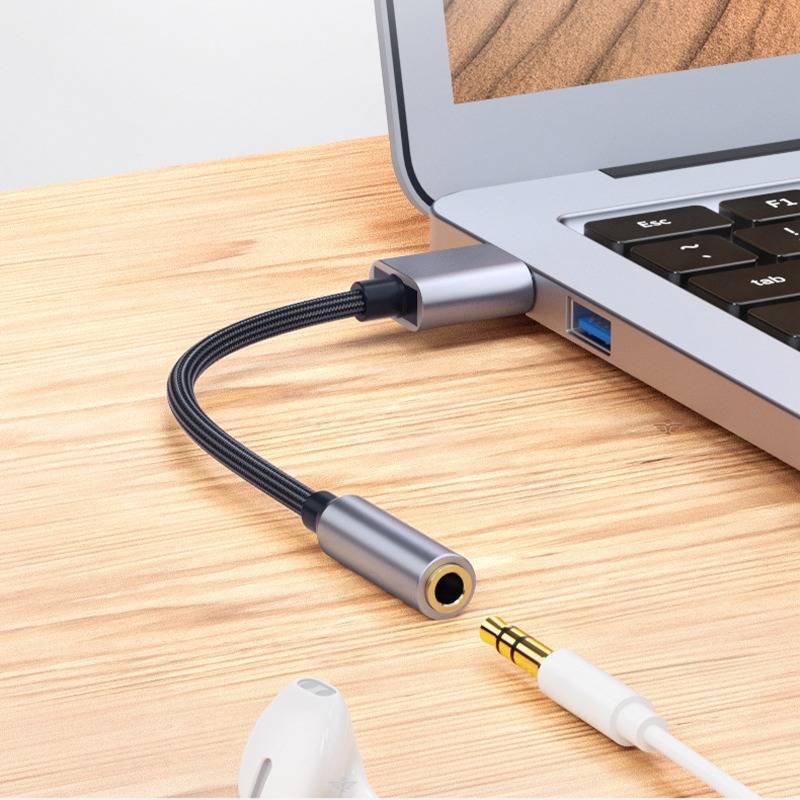 Laptop, der über ein Audioadapter mit 3,5-mm-Kopfhörerklinken-Kabel und USB-Stecker auf einem Holzschreibtisch angeschlossen ist.