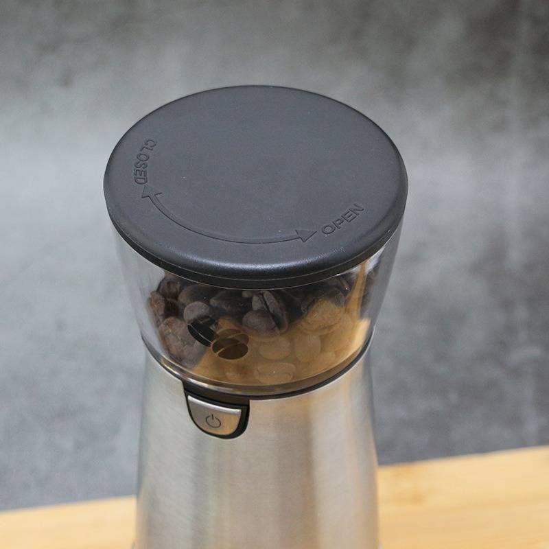 Elektrische Kaffeemühle mit Bohnen im Inneren, gezeigt auf einer Holzoberfläche vor einem grauen Hintergrund.