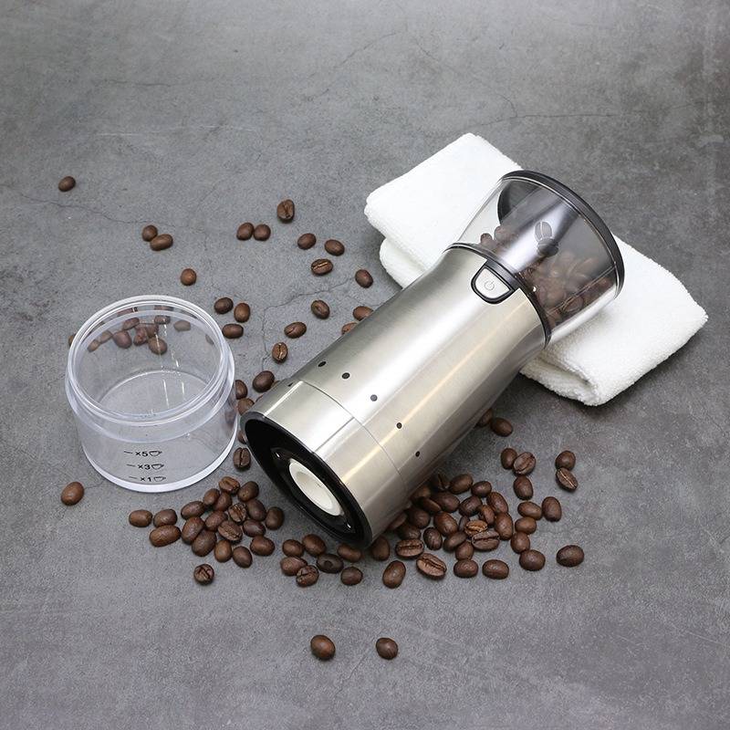 Elektrische Kaffeemühle umgestürzt auf ihrer Seite neben einer Messbechern, verschüttete Kaffeebohnen und ein gefaltetes weißes Handtuch auf einer grauen Oberfläche.