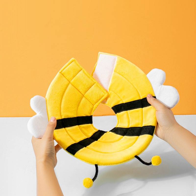 Hände halten ein gelbes bienenförmiges Plüsch-Ringspielzeug mit schwarzen Streifen, weißen Flügeln und baumelnden Pom-Pom-Füßen auf orangeweißem Hintergrund.