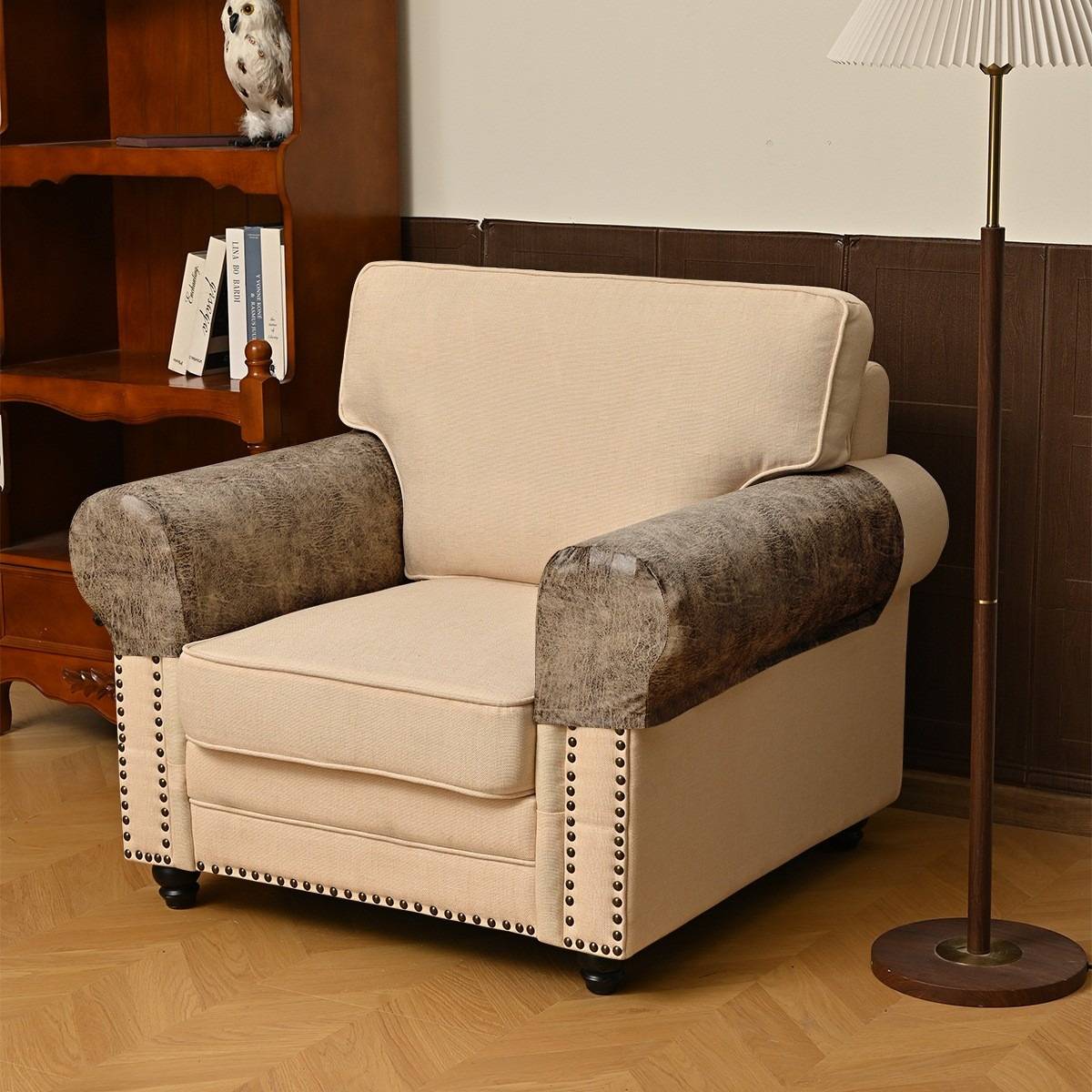 Ein beiger, gepolsterter Sessel mit grauen Armlehnen sitzt auf einem Holzfußboden neben einer Stehleuchte und einem Bücherregal in einem Wohnzimmer.