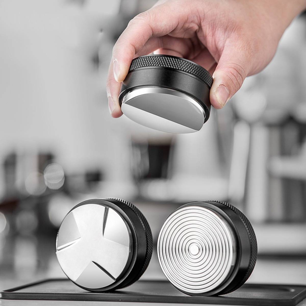 Hand, die ein kreisförmiges Espresso-Tamper-Werkzeug über zwei Metall-Kaffee-Zubehörteilen auf einem Tablett in einer Küchen-Einstellung platziert.