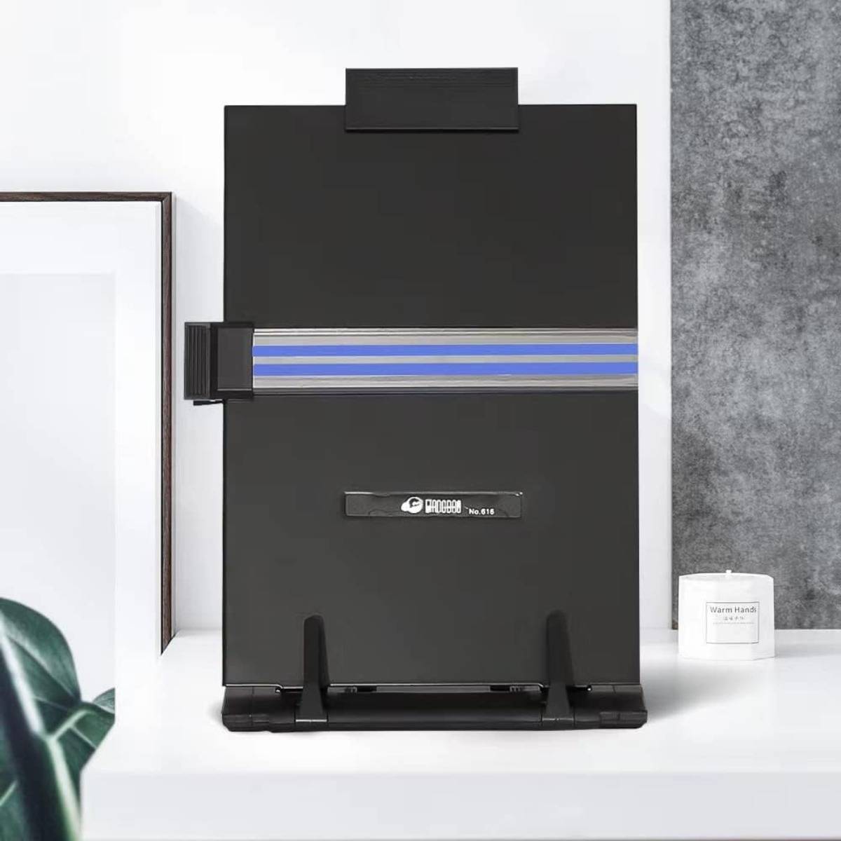 Ein schwarzes Falttafel- oder Staffeleibrett mit einem horizontalen blaugrauen Streifen steht auf einem weißen Regal neben gerahmter Kunst und einer Kerze.