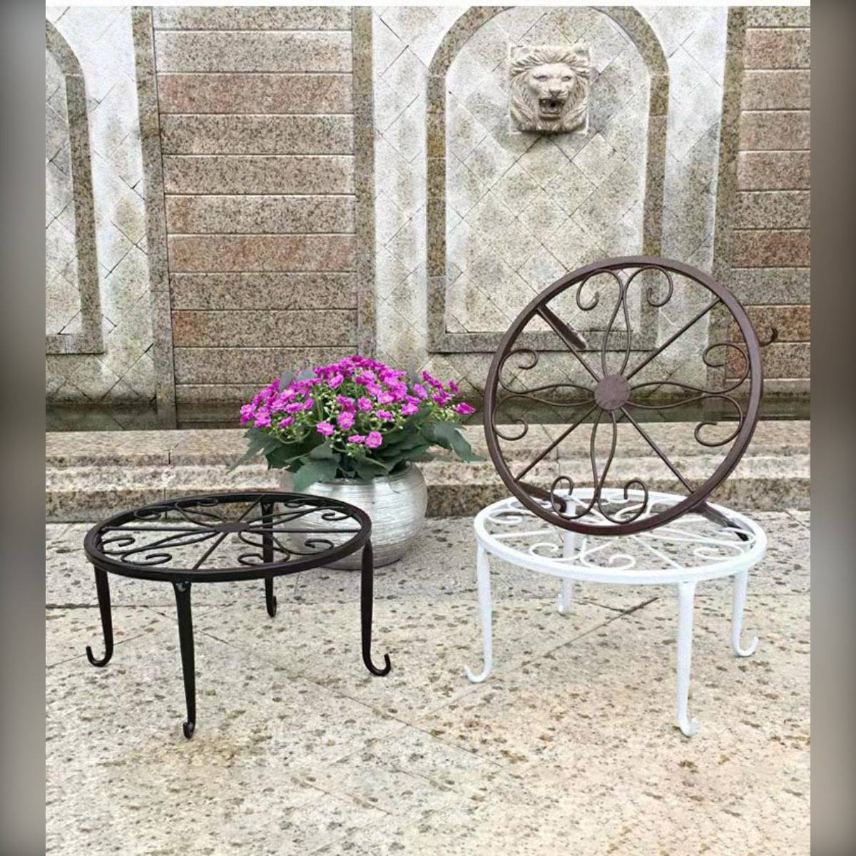 Zwei dekorative Metall-Pflanzständer oder Tische auf einer Steinterrasse, einer schwarz und einer weiß mit einem Wagenrad-Design, neben einem Topf mit rosa Blumen.