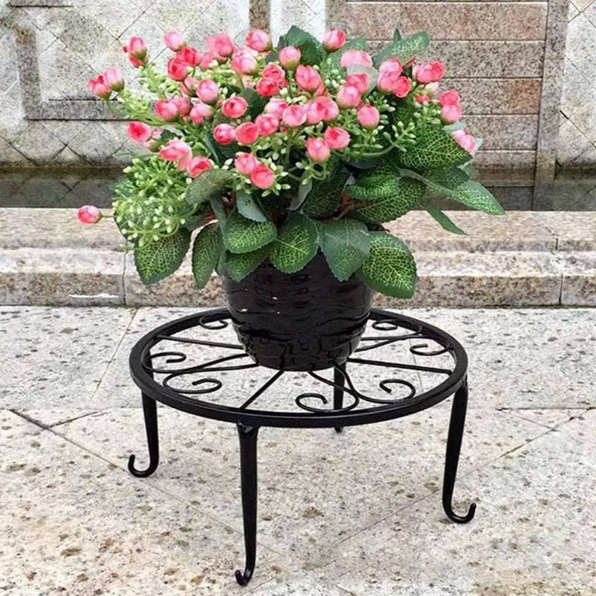 Getopfte rosa blühende Pflanze in einer schwarzen Vase auf einem dekorativen Metallständer im Freien auf Steinpflaster.