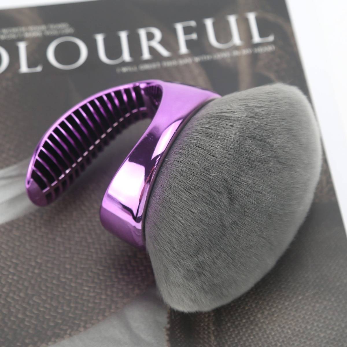 Nahaufnahme eines flauschigen Makeup-Pinsels mit glänzendem violettem Griff auf einer dunklen Magazin- oder Produktseite.